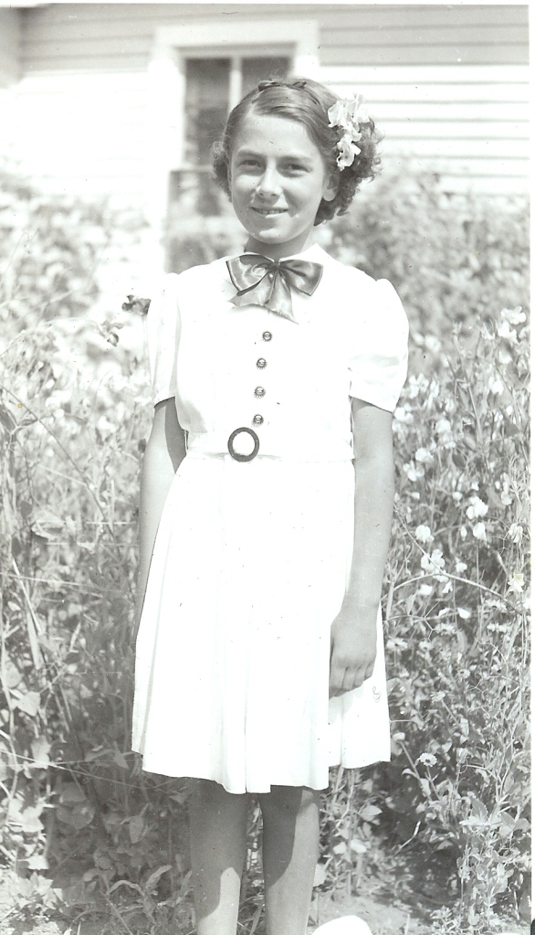 Image of Sylvia Fedoruk at age 12, dressed in a white dress with a neck bow and wearing flowers in her hair, ready to greet royalty in 1939.
